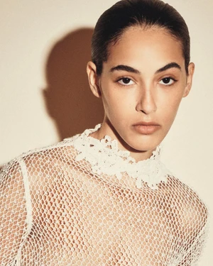 Close-up portrait of a model wearing a white lace mesh top against a warm studio background with a sharp shadow.