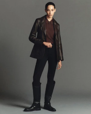 Full-length portrait of a model wearing a brown leather jacket, brown polo shirt, black trousers, and tall black boots against a grey studio backdrop