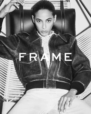 Model wearing a leather bomber jacket seated in a woven strap chair with FRAME logo overlaid across the chest