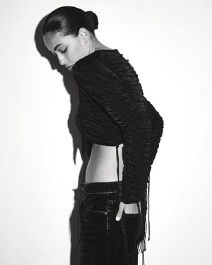 Model in a black long-sleeve crop top and dark jeans posing against a white wall with strong shadow cast.