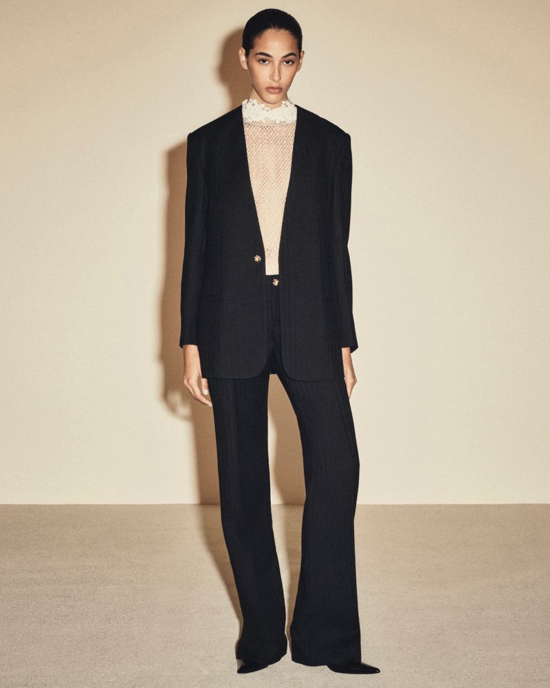 Full-length portrait of a model wearing a black blazer and trousers with a cream lace-collared top against a neutral studio backdrop.