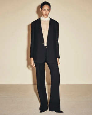 Full-length portrait of a model wearing a black blazer and trousers with a cream lace-collared top against a neutral studio backdrop.