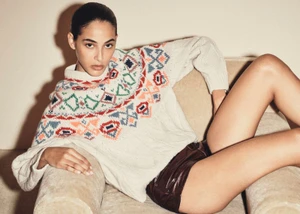 Model reclining in a cream patterned sweater and dark shorts on an upholstered armchair against a plain wall.