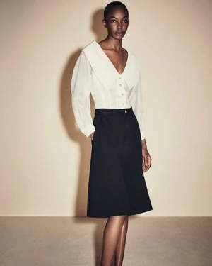 Model wearing a white collared blouse and black skirt against a neutral studio backdrop