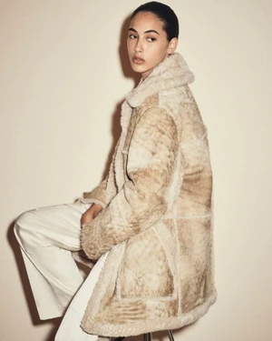 Model in textured shearling coat and white trousers seated against a neutral studio background.