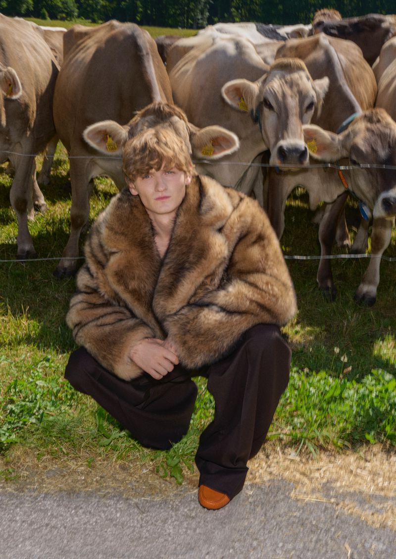 A young person in a fur coat poses in front of a herd of cows on a grassy field, wearing brown trousers and a brown shoe.