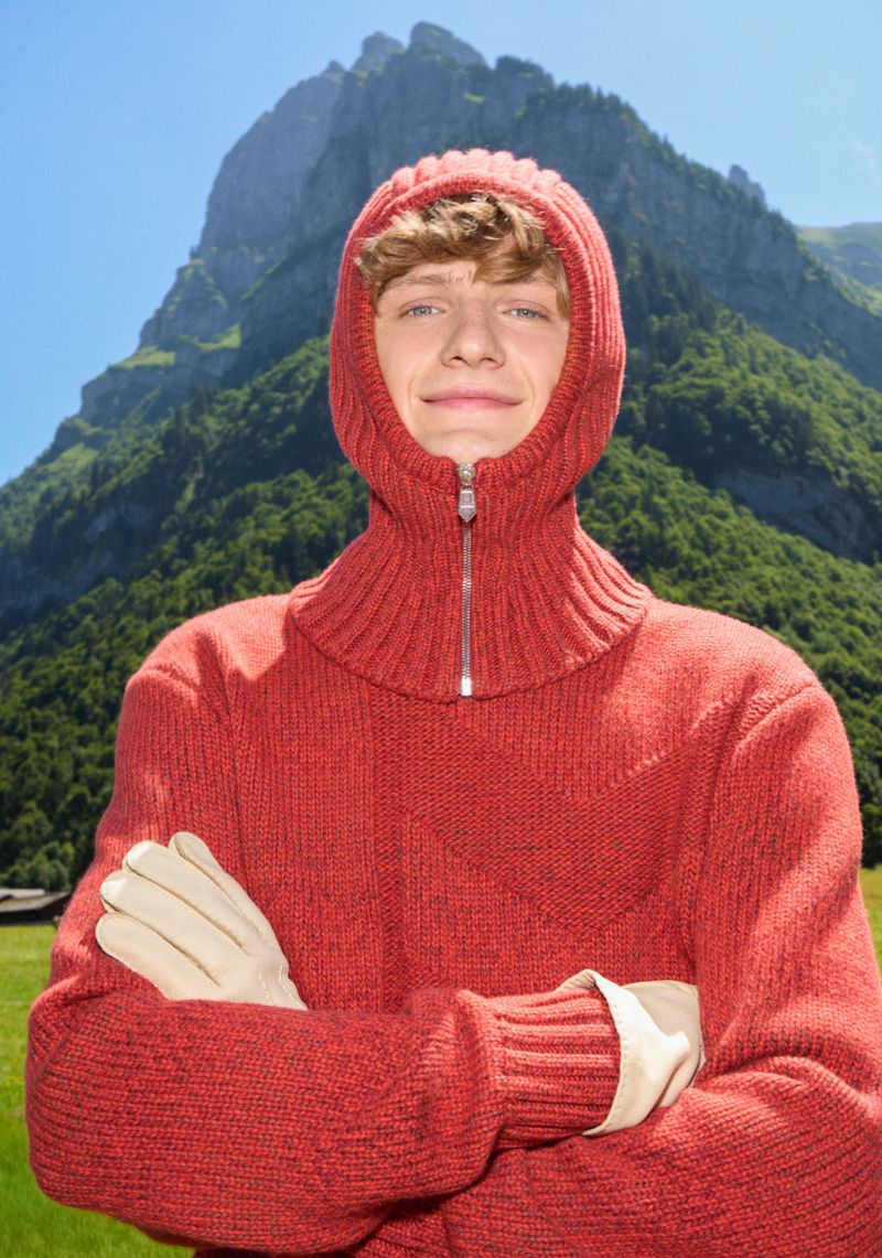 A young adult with red hair stands outdoors in a hooded red sweater and gloves against a backdrop of mountains and green fields.