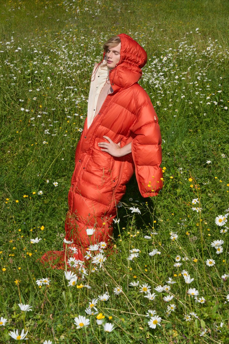 Blonde-haired person wearing a red hooded puffer sleeping bag suit in a field of daisies on a sunny day, creating a whimsical outdoor fashion shot.