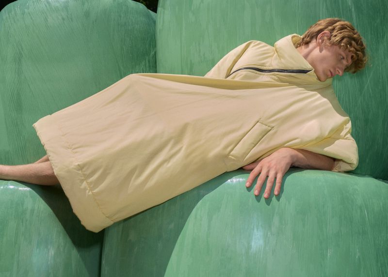 Adult male model with light hair lying on green wrapped hay bales while wearing a large light-yellow jacket in an outdoor setting.