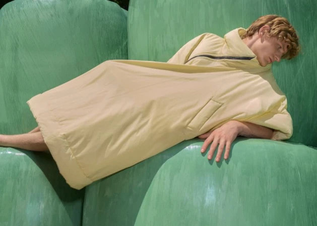 Adult male model with light hair lying on green wrapped hay bales while wearing a large light-yellow jacket in an outdoor setting.