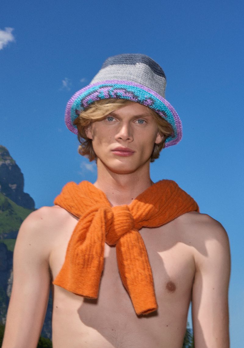 Man with an orange sweater tied around his torso, wearing a crochet bucket hat, posing in front of a mountain backdrop against a blue sky.