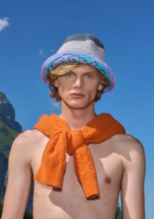 Man with an orange sweater tied around his torso, wearing a crochet bucket hat, posing in front of a mountain backdrop against a blue sky.