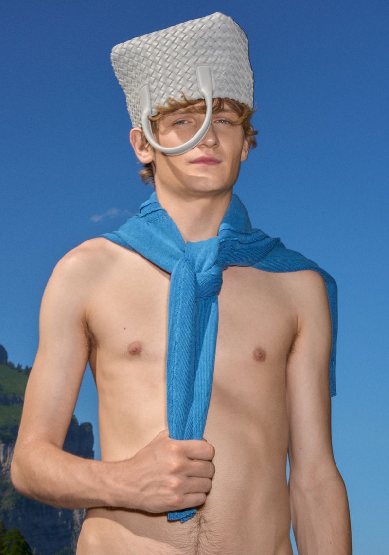 Adult male poses with a woven white hat and a blue scarf draped around his shoulders against a bright blue sky with green mountain backdrop.