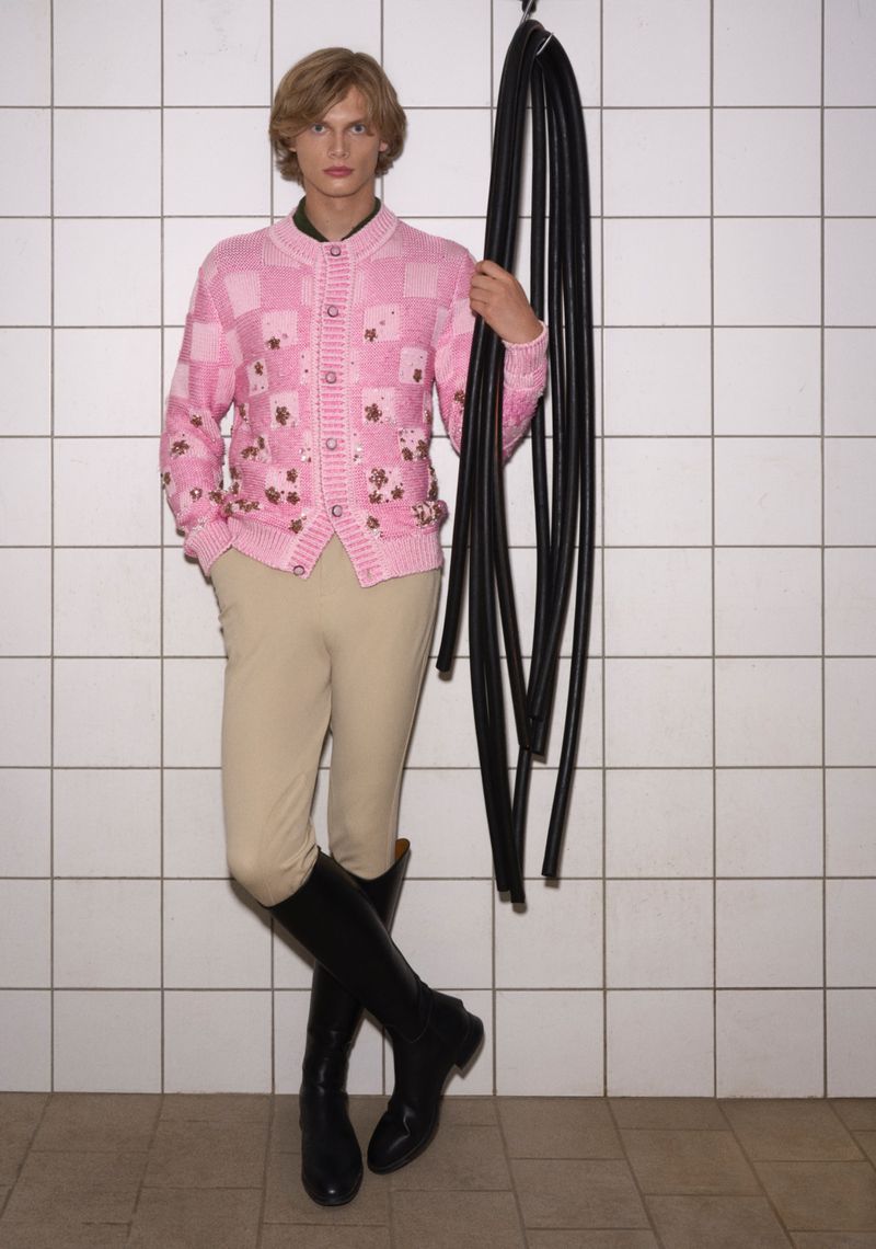 Man in a pink patterned cardigan, beige jodhpurs, and black boots stands against a tiled wall holding black hoses on a hook.