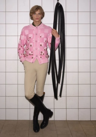 Man in a pink patterned cardigan, beige jodhpurs, and black boots stands against a tiled wall holding black hoses on a hook.