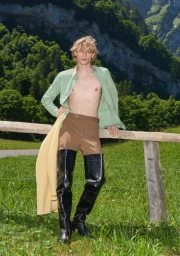 Blonde man wearing an unbuttoned sage green shirt, brown trousers, and tall black boots leaning on a wooden fence outdoors.