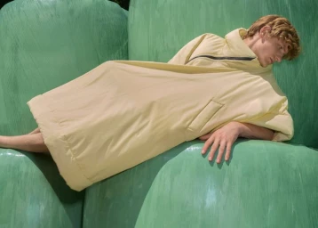 A male model rests in an oversized pale yellow padded garment across glossy green sculptural curved shapes.
