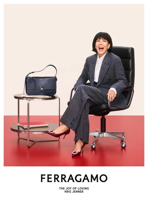 Kris Jenner laughing while seated in a black office chair wearing a grey tweed suit and burgundy heels for Ferragamo Holiday 2026