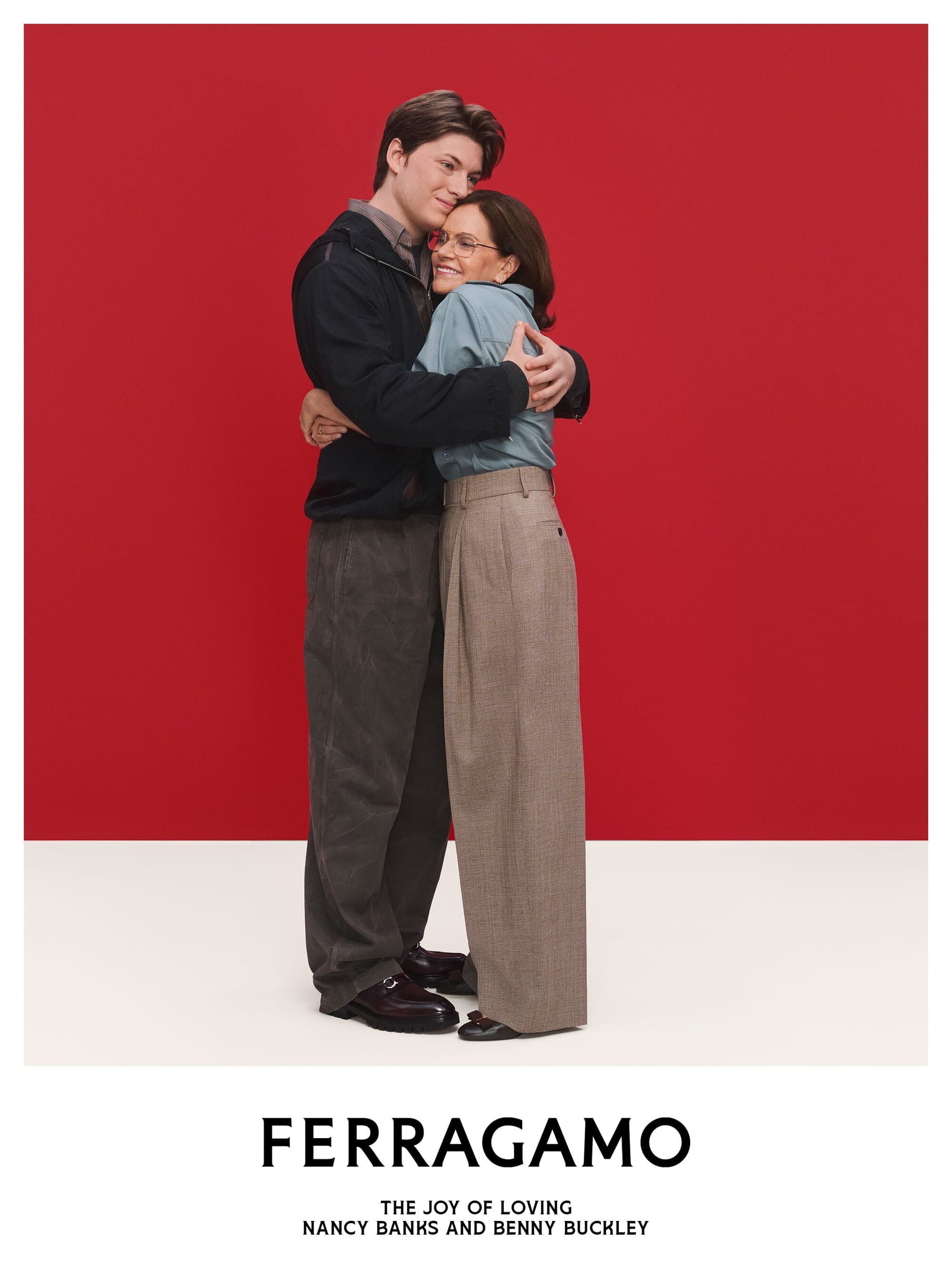 Two people embracing against a solid red backdrop with a white floor featuring the Ferragamo logo and text below.