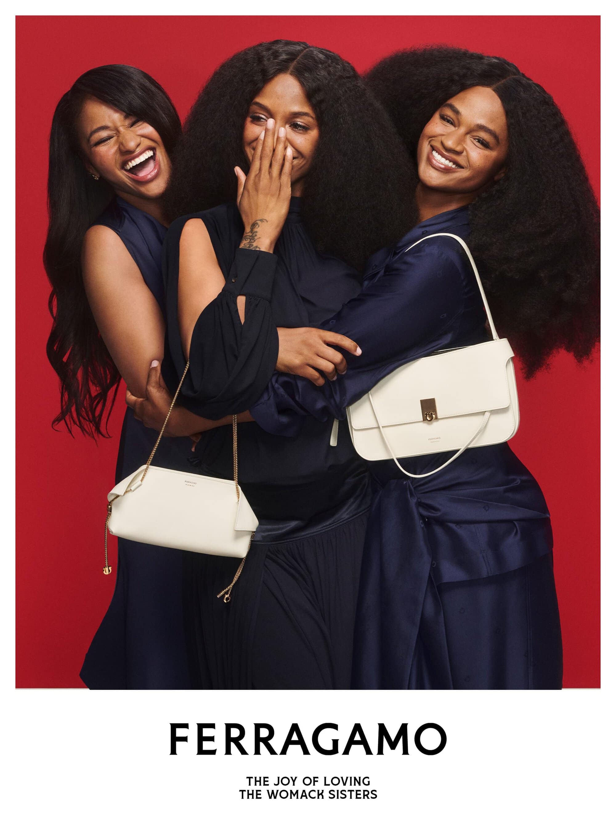 Three smiling women in dark navy blue dresses holding white leather handbags against a solid red background