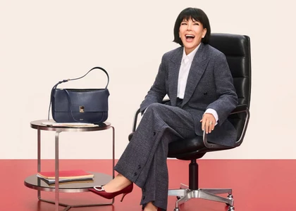 Kris Jenner laughing while seated in a black office chair wearing a grey tweed suit and burgundy heels for Ferragamo Holiday 2026