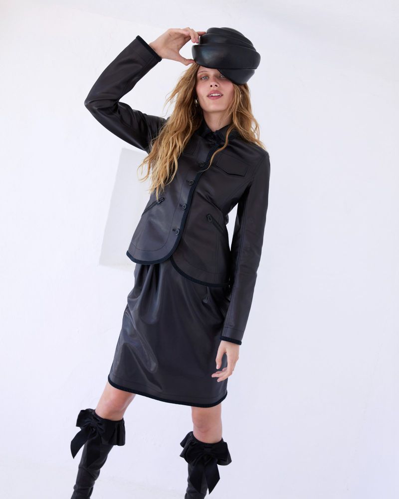 A female fashion model poses in a dark leather jacket, skirt, and hat against a white backdrop in a studio setting with bright lighting.