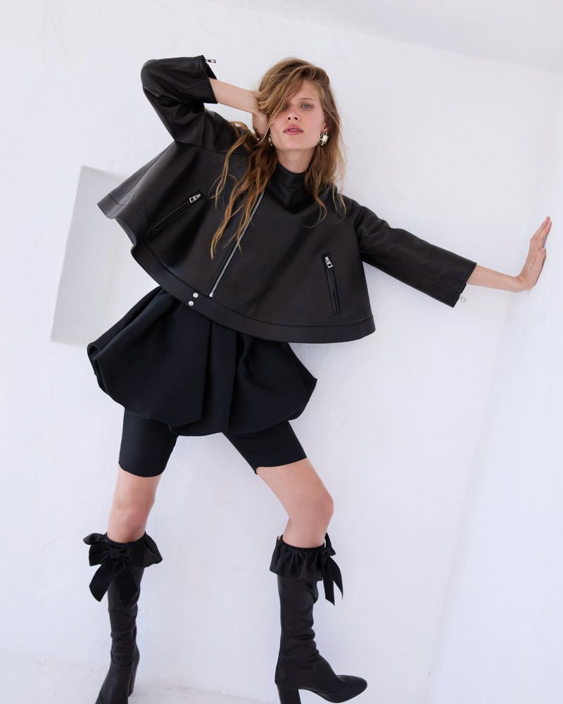 Fashion shot of a stylish adult female model in a black leather jacket, skirt, and knee-high boots posing against a white backdrop.