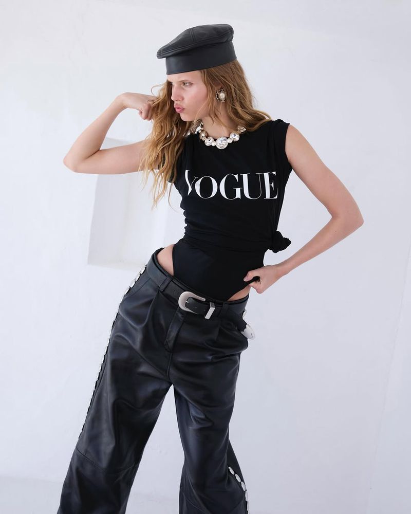 Fashionable woman strikes a pose in black top with Vogue logo, black leather beret, pearl necklace, and baggy leather pants on white.