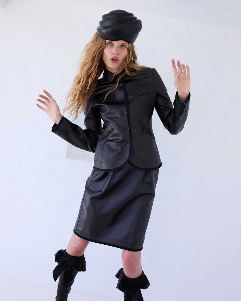 Fashion model poses in a black leather skirt suit with a matching hat against a white background in a studio setting for a portrait.