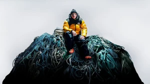 Man in yellow puffer jacket seated atop a massive pile of tangled climbing ropes against a plain white studio backdrop.