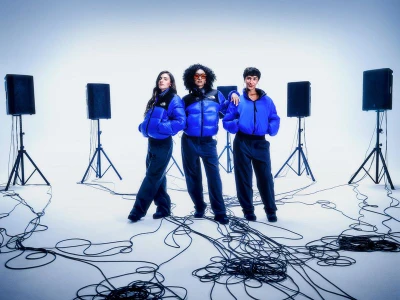 Three models wearing blue puffer jackets and dark trousers pose on a white studio floor surrounded by speaker stands and tangled cables.