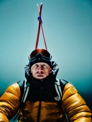 Close-up portrait of a male mountaineer wearing a yellow down puffer jacket, black beanie, and mirrored ski goggles with skis upright behind him