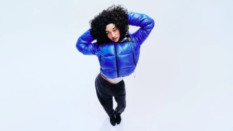 Woman with curly dark hair wearing a bright blue puffer jacket and dark trousers against a white background