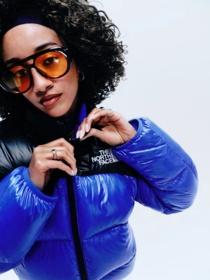Model with tinted sunglasses wearing a blue and black puffer jacket against a white background