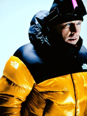 Man wearing a yellow and black puffer jacket, black beanie, and ski goggles against a bright white background.