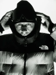 Close-up of a person in a puffer jacket and ski goggles adjusting the hood against a plain white background