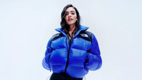 Woman wearing a bright blue puffer jacket with black panels against a plain white studio background.