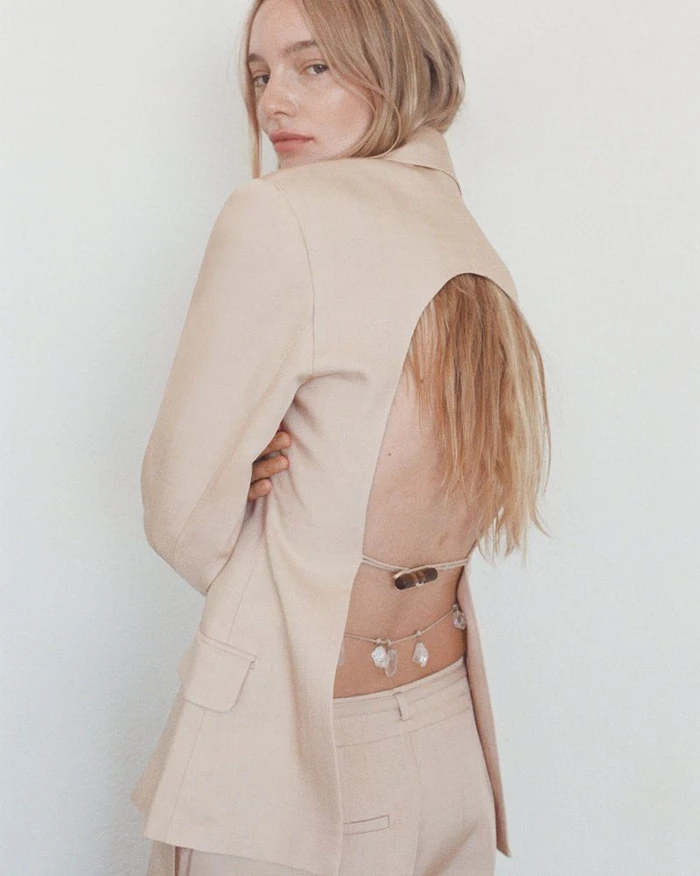 Model Maya Stepper wearing a beige open-back blazer and trousers against a white wall.
