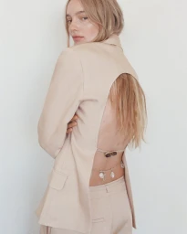 Model Maya Stepper wearing a beige open-back blazer and trousers against a white wall.