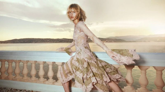 Model Aivita Muze wearing a patterned dress posing on a balcony overlooking water at sunset.