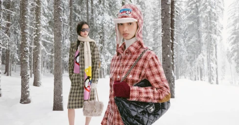 Two models in winter attire standing in a snowy forest for Chanel Coco Neige, featuring a red tweed hat, jacket, and black quilted bag.
