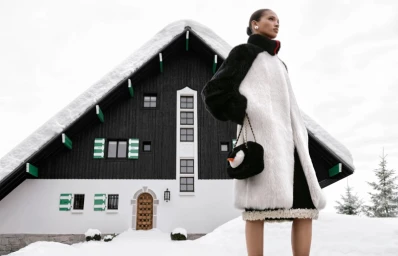 Model in a black and white faux fur coat standing in front of a snow-covered chalet for Chanel Coco Neige 2026