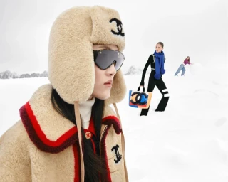 Model in foreground wearing a beige shearling ushanka hat and matching coat with dark sunglasses against a snowy winter background.