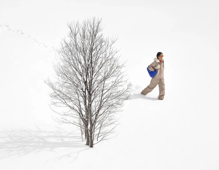 Model walking through deep snow past a bare winter tree, wearing a patterned ski suit and carrying a blue bag for Chanel.