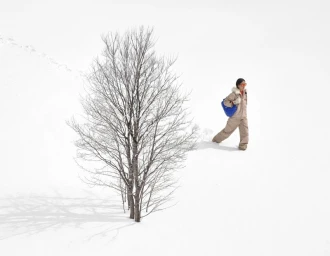 Model walking through deep snow past a bare winter tree, wearing a patterned ski suit and carrying a blue bag for Chanel.