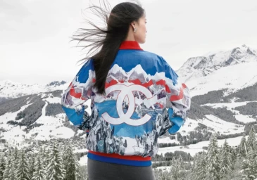 Model seen from behind wearing a blue ski jacket with mountain graphic and Chanel logo against a snowy mountain backdrop.