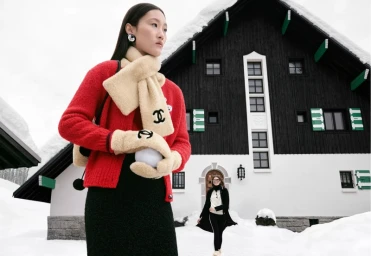 Model in red cardigan and cream shearling scarf standing in the snow in front of a chalet for Chanel Coco Neige 2026