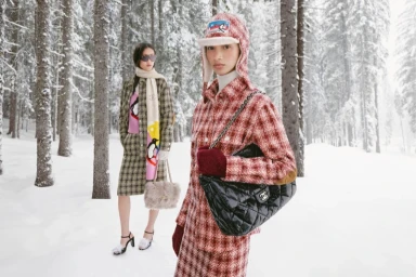 Two models in winter tweed apparel and puffer accessories posing outdoors in a snowy pine forest.