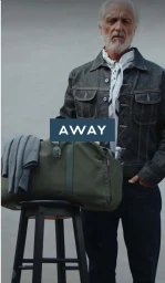 Elderly man in denim jacket standing beside a green weekender bag resting on a stool for AWAY Travel