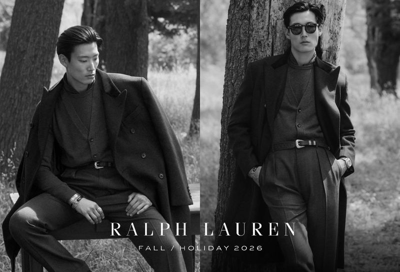 Black and white fashion campaign image featuring two male models in tailored coats and trousers outdoors near trees
